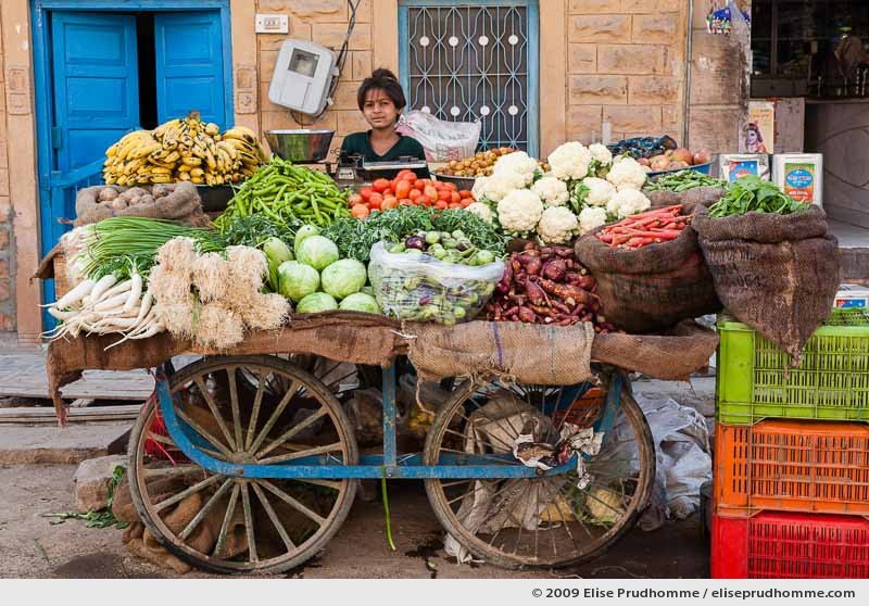 A young girl standing behind a fruit and vegetable trolley in Rohet village, Rajasthan, Jodhpur, India, 2009 by Elise Prudhomme.