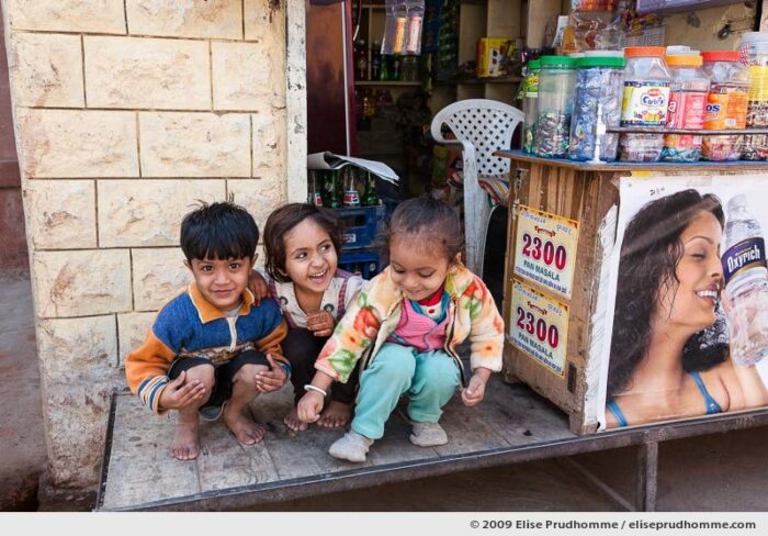 Three smiling children in front of a small store, Oisan, Rajasthan, India, 2009 by Elise Prudhomme.