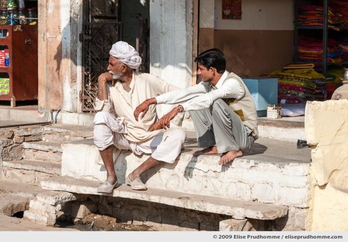 Two Indian men, one wearing traditional clothing, sitting in front of a shop in Rohet village, Rajasthan, Jodphur, India, 2009 by Elise Prudhomme.