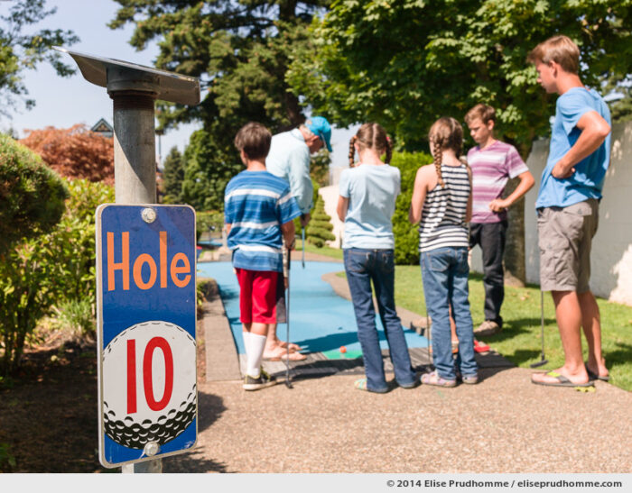 A grandfather and his grandchildren gather at the tenth hole of Wilsonville Family Fun Center miniature golf course on a sunny clear summer day, Oregon, USA. Focus on foreground and panel of the 10th hole. Un grandpère et ses petits-enfants se rassemblent au dixième trou du parcours de Golf Miniature par un jour d'été clair et ensoleillé, Wilsonville, Oregon, USA. Mise au point sur le panneau en avant plan.