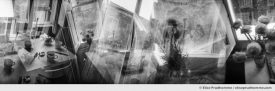 Black and white panoramic photograph of louvered blinds, ranunculus bouquet and tea for two in a Roman apartment, Italy. Analog photography series entitled Lieux-dits by Elise Prudhomme.
