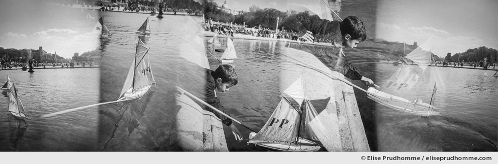 Black and white panoramic photograph of a boy sailing his boat in the basin at Luxembourg Garden, Paris, France. Analog photography series entitled Lieux-dits by Elise Prudhomme.