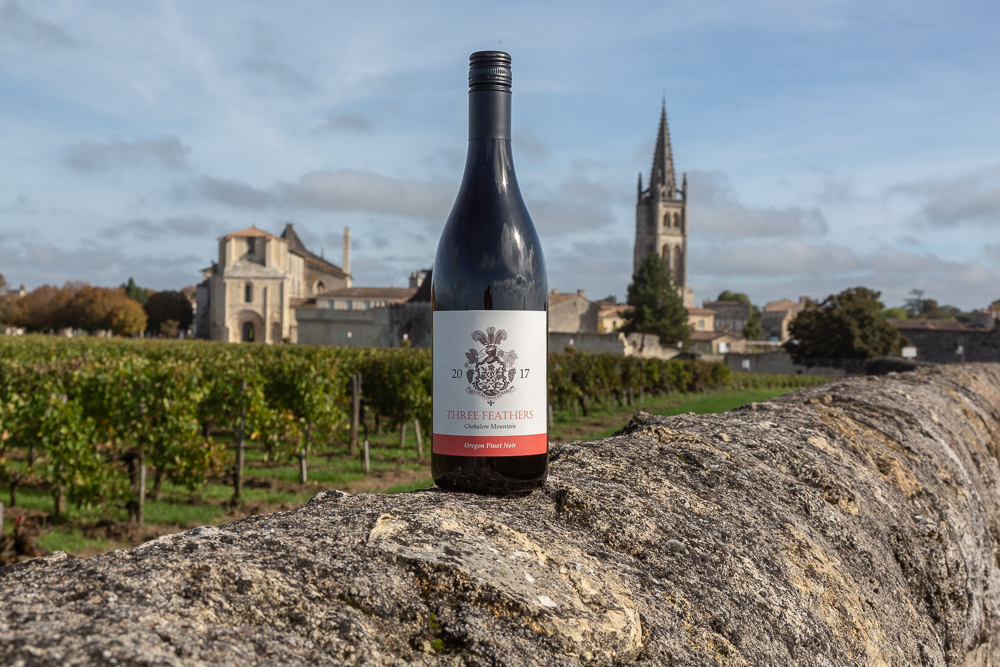 A bottle of Three Feathers Estate Pinot Noir perched on a vineyard wall with the town Saint-Emilion in the background, Bordeaux region, Gironde, France.